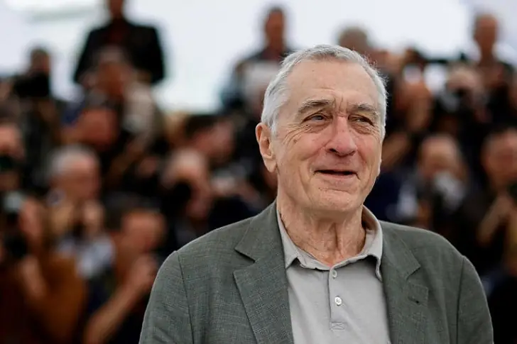 Net worth of Robert De Niro - Robert De Niro with a warm smile and grey hair dressed in a grey blazer over a casual shirt.